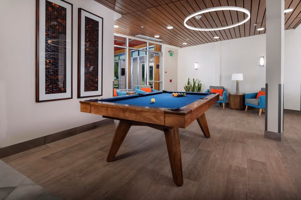 A modern indoor recreational room featuring a wooden pool table with a blue felt surface and billiard balls arranged on it. The room has a wooden ceiling with circular light fixtures, light-colored walls, and wood-look flooring. In the background, there are two blue armchairs with orange cushions, a small round wooden table with a lamp, and some green plants near a glass door leading outside. Two framed abstract artworks hang on the wall to the left.