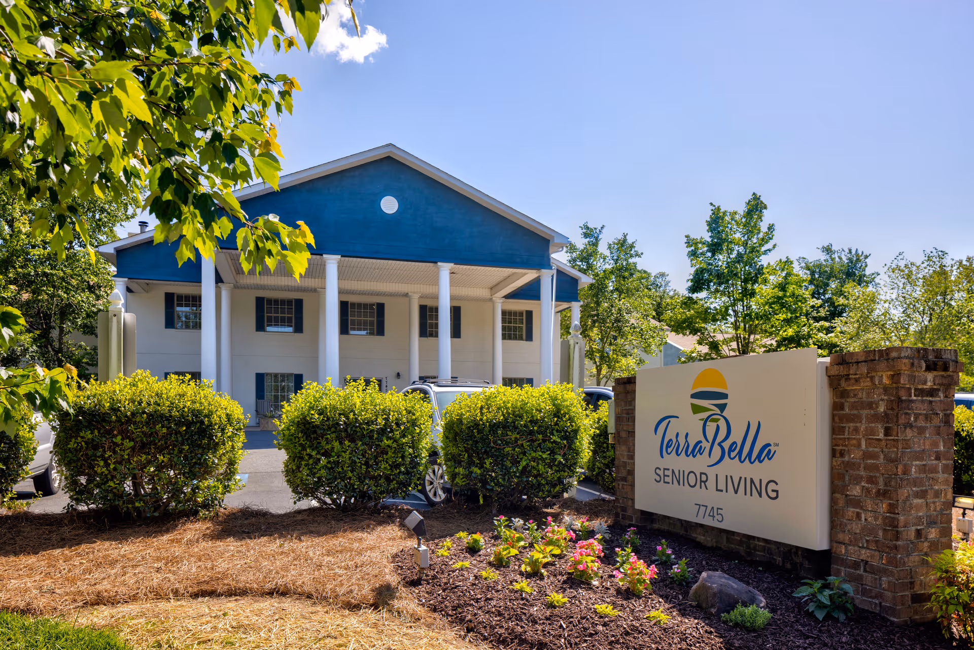 Exterior view of TerraBella Senior Living facility with a blue and white building featuring large white columns, surrounded by green bushes and trees under a clear blue sky. A sign in front reads 'TerraBella Senior Living 7745' with a colorful logo above the text.