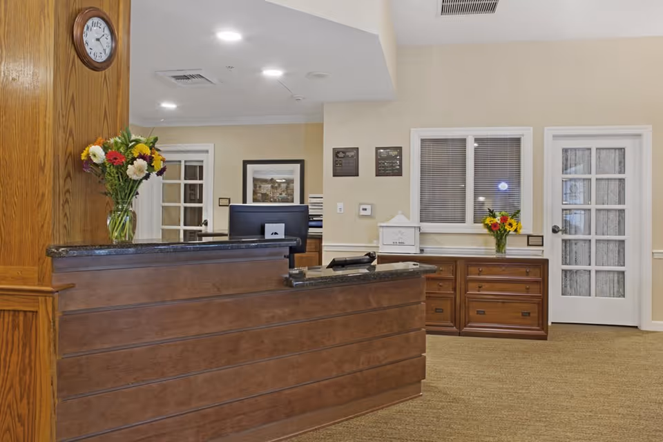 Reception area with a wooden front desk featuring a granite countertop, a vase of colorful flowers on the desk, a clock on the wooden wall, and a cabinet with another vase of flowers. The background shows a computer monitor, framed artwork, plaques on the wall, a window with blinds, and two doors with glass panels.
