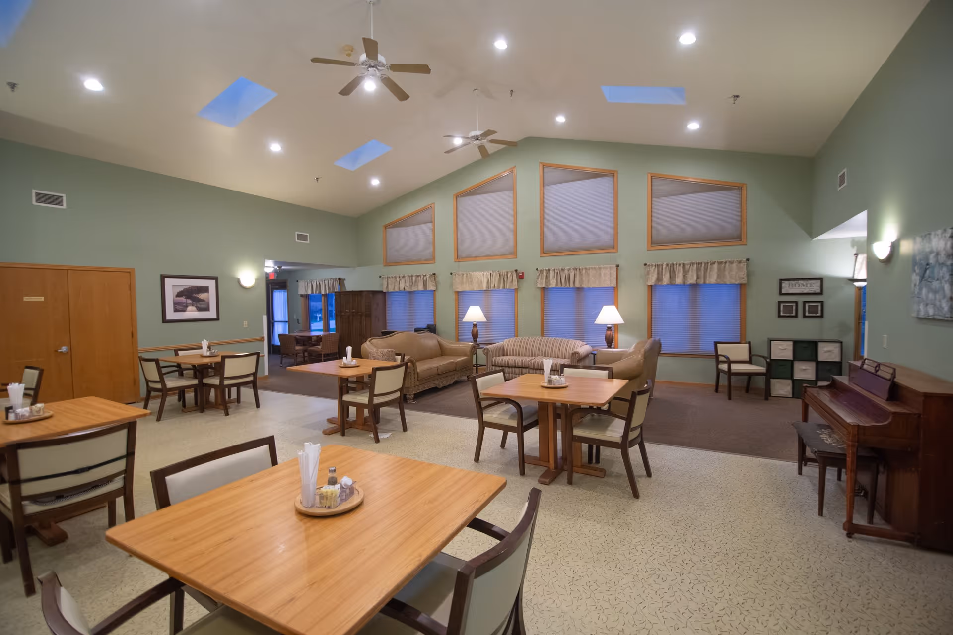 Spacious communal dining and lounge area with wooden tables and chairs, sofas, lamps, and a vaulted ceiling with skylights and ceiling fans.