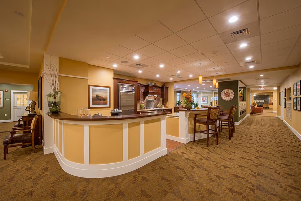 Interior view of a senior living facility featuring a curved reception desk with decorative vases and a framed picture on the wall. The area has warm yellow walls, patterned carpet, and recessed lighting. There are high chairs at a counter, a large clock on a green accent wall, and a hallway leading to other rooms with seating areas visible in the distance.