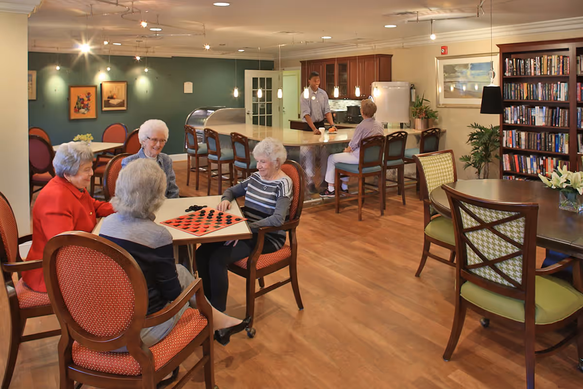 A group of elderly women sitting around a table playing checkers in a well-lit common area with wooden floors. In the background, a kitchen area with a man and a woman sitting at the counter is visible. There is a bookshelf filled with books and a round table with chairs on the right side of the room.