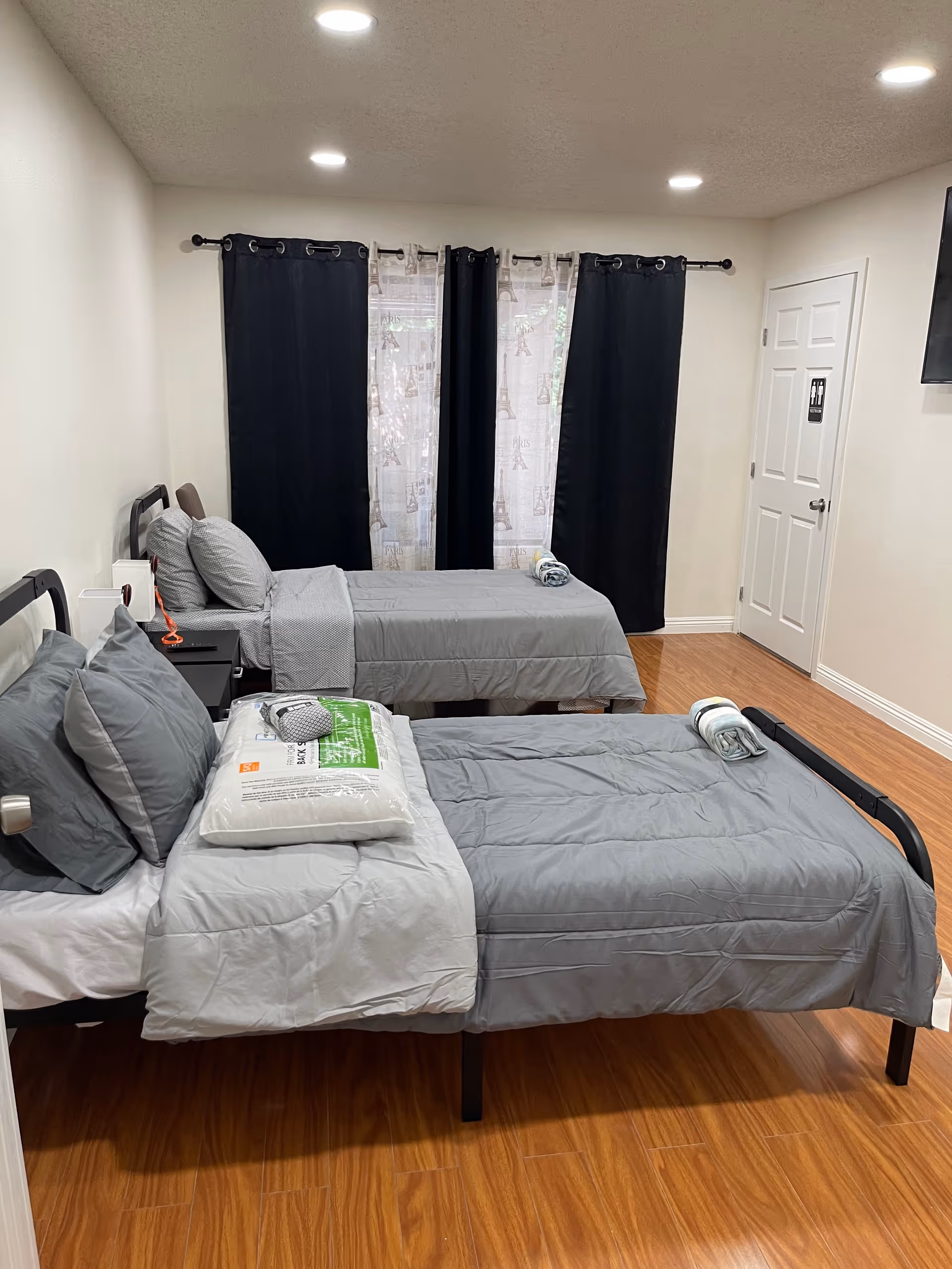 A shared bedroom in a senior living facility with two single beds, gray bedding, and dark curtains.