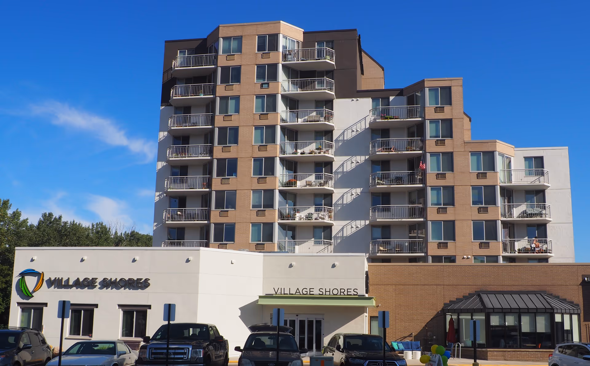 Exterior view of Village Shores, a multi-story senior living facility with balconies on each floor, a clear blue sky, and several parked cars in front of the building.