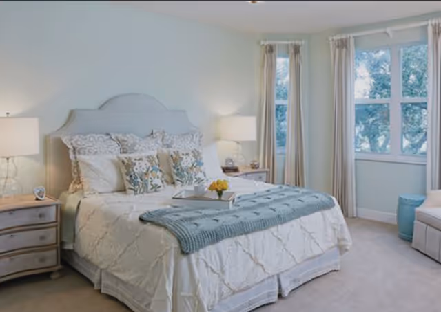 A bright and cozy bedroom with a large bed featuring a light-colored upholstered headboard, white bedding, decorative pillows, and a blue throw blanket. There are two wooden nightstands with lamps on either side of the bed. The room has light-colored walls, carpeted floor, and large windows with beige curtains letting in natural light. A small blue stool and a cushioned chair are placed near the windows.
