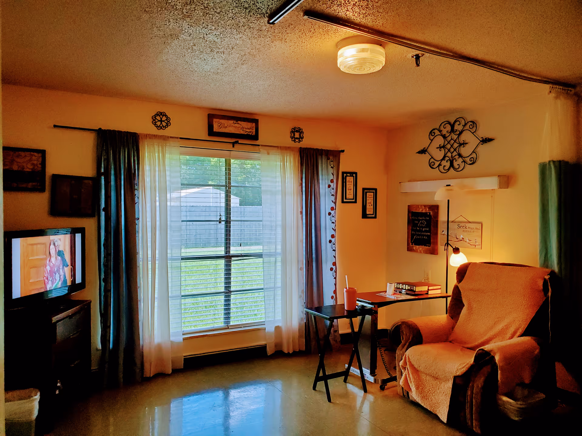 A cozy room with a large window covered by sheer and dark curtains, a comfortable armchair draped with a beige cover, a small table with a drink cup, a floor lamp, and a television on a stand showing a woman. The walls are decorated with framed pictures and ornamental wall hangings.