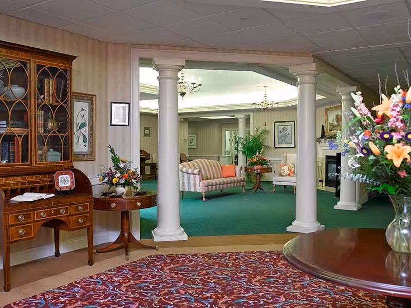 A spacious and elegantly decorated living area in a senior living facility featuring green carpet, white columns, a floral patterned rug, wooden furniture including a cabinet and tables with flower arrangements, and comfortable seating with striped and floral upholstery.