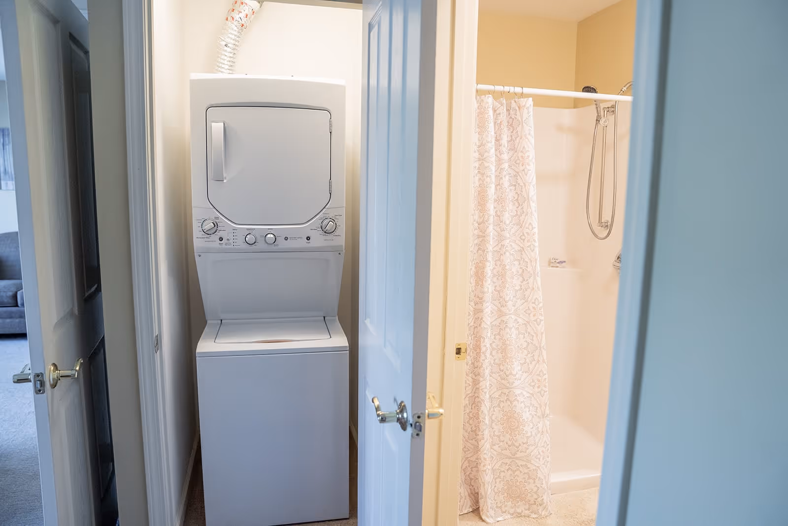 A small laundry area with a stacked washer and dryer unit next to a bathroom with a shower and a patterned shower curtain, viewed through partially open doors.
