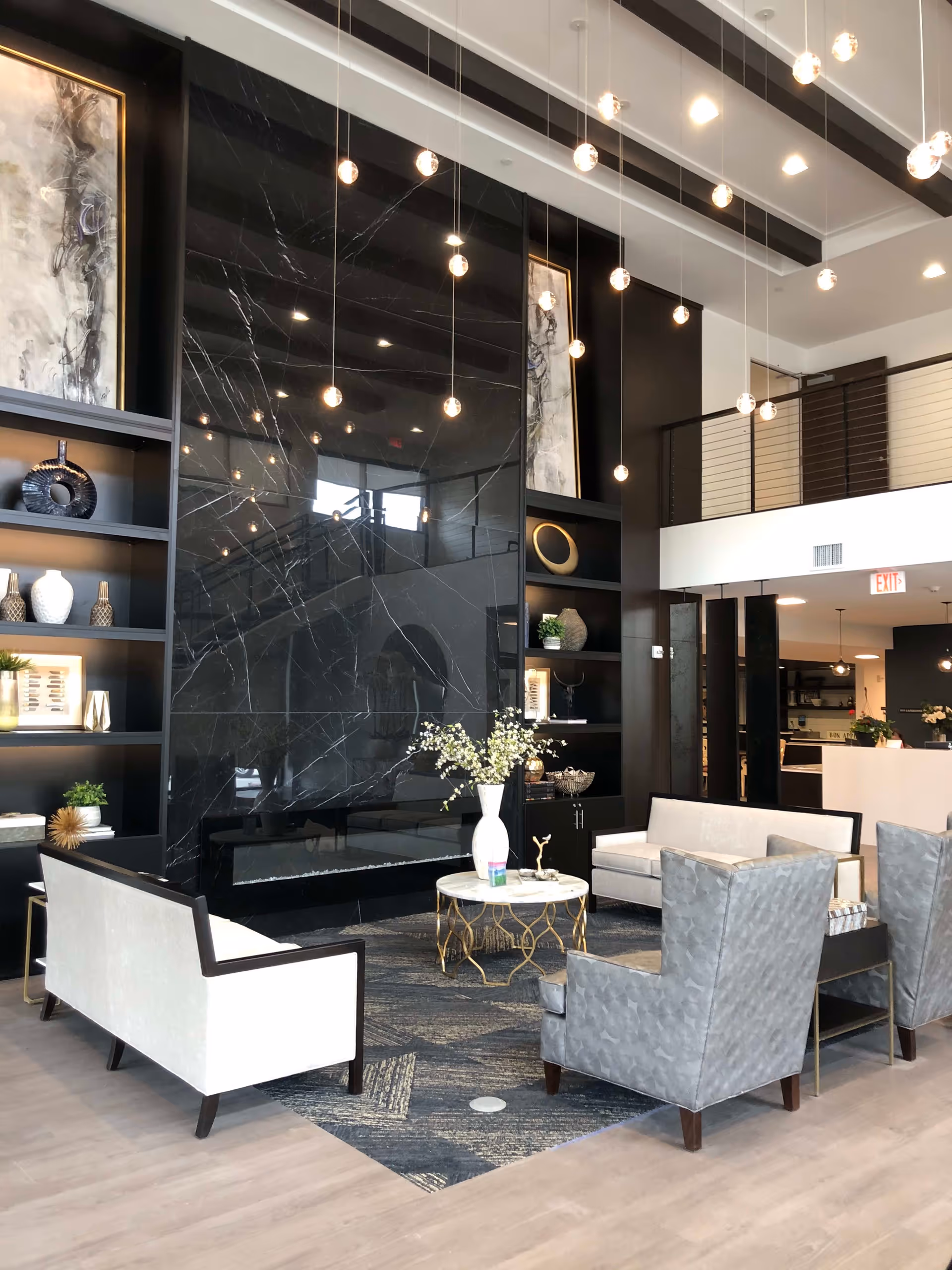 A modern and elegant living room area with a large black marble fireplace as the focal point. The room features two white sofas, two gray armchairs, a round marble coffee table with a white vase and flowers, and built-in black shelves with decorative items. Pendant lights hang from the ceiling, and there is a mezzanine level with a railing visible in the background.