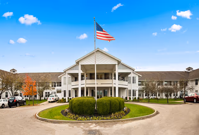 Front exterior view of a large white senior living facility building with a circular driveway and neatly trimmed bushes in the center. An American flag is flying on a flagpole in front of the building under a bright blue sky with a few clouds.