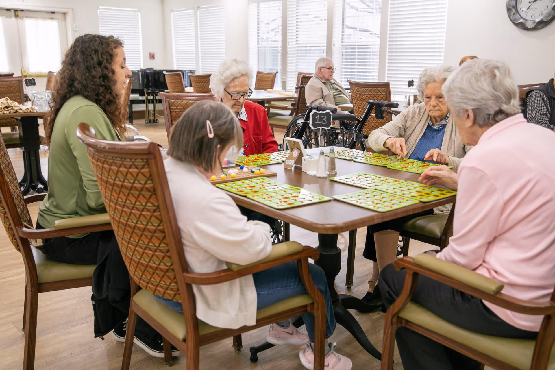 A group of elderly people and a caregiver sitting around a table playing bingo in a bright, spacious room with large windows and wooden flooring.
