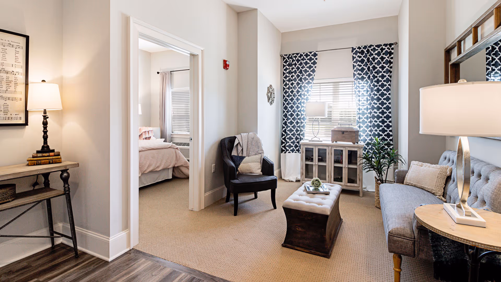 A cozy living room area with a gray sofa, a black armchair with pillows, a tufted ottoman, and a wooden side table with a lamp. The room has beige carpet and light-colored walls. A window with patterned curtains lets in natural light. Through an open doorway, a bedroom with a bed and window blinds is visible.