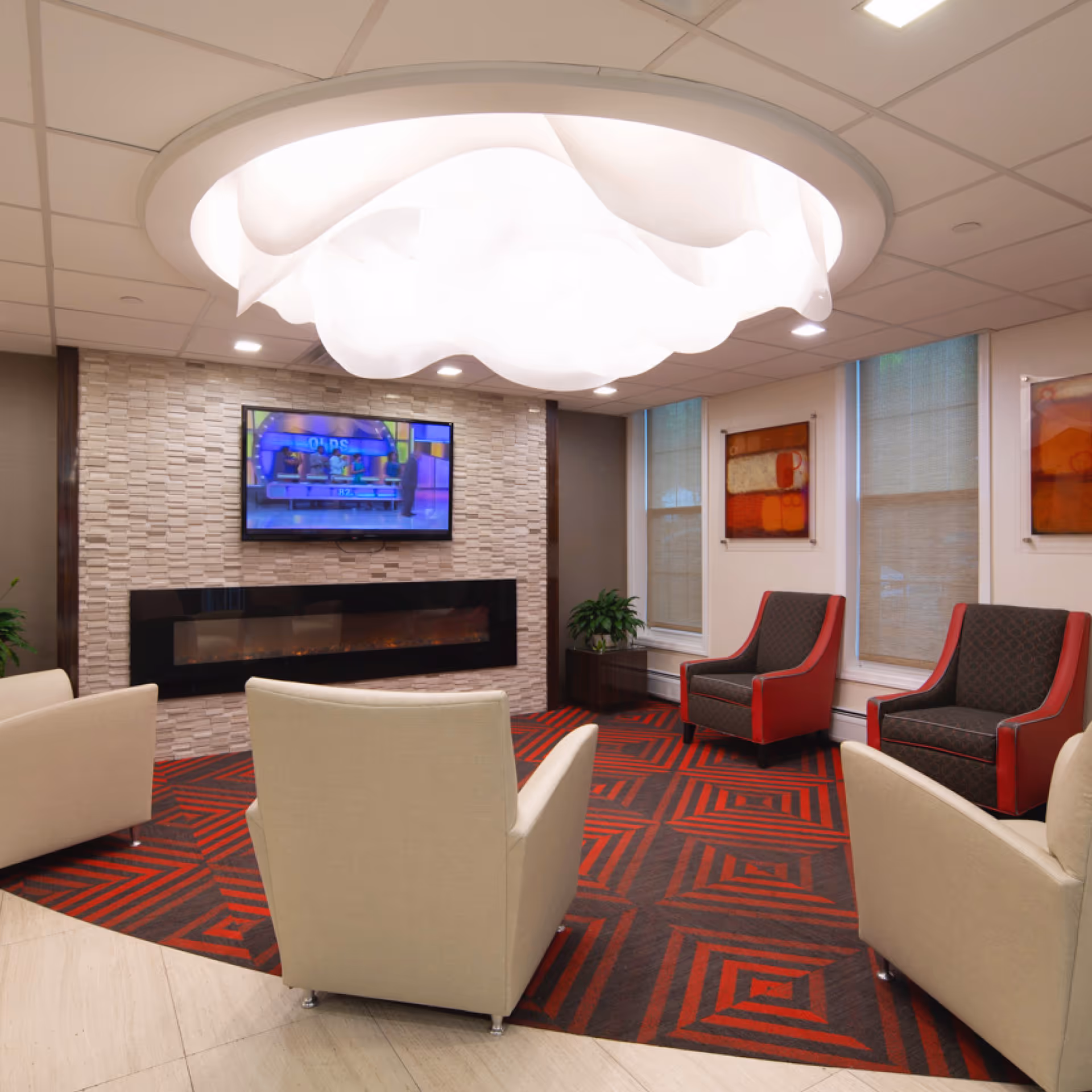 A modern living room area with a large, artistic ceiling light fixture, a wall-mounted flat-screen TV above a sleek electric fireplace, beige and red armchairs arranged on a red and black patterned carpet, two windows with blinds, and abstract artwork on the walls.