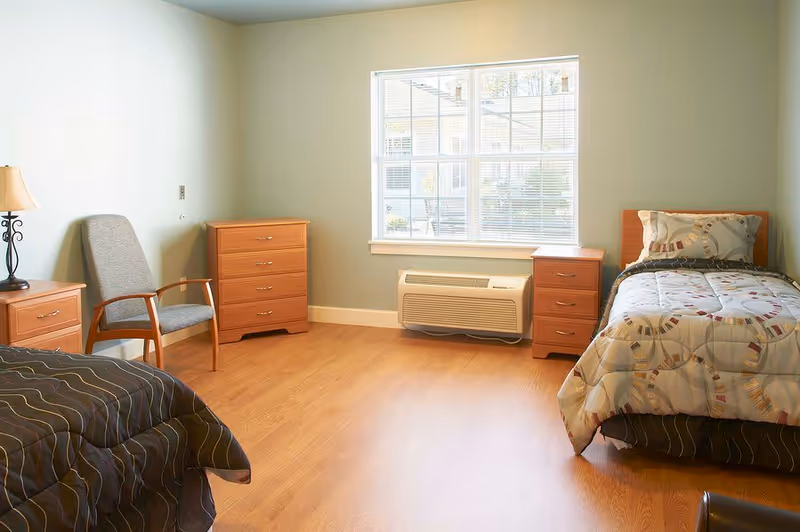 A bright and clean bedroom in an assisted living facility with two single beds, each with patterned bedding. The room has light green walls, a large window with blinds, wooden flooring, a gray upholstered chair, and wooden furniture including two nightstands and a chest of drawers. An air conditioning unit is installed below the window.