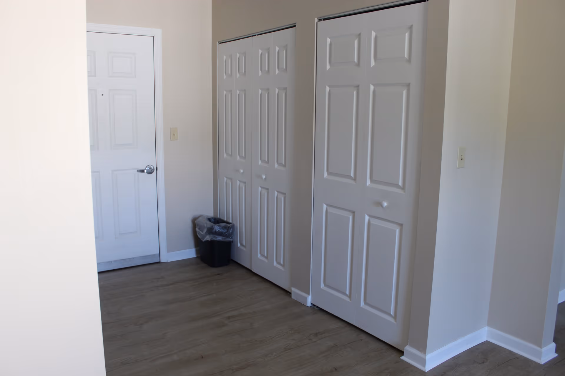 Interior view of a hallway area with light-colored walls and wood flooring. There is a white door on the left side, two sets of white closet doors on the right, and a small black trash bin placed near the closets.