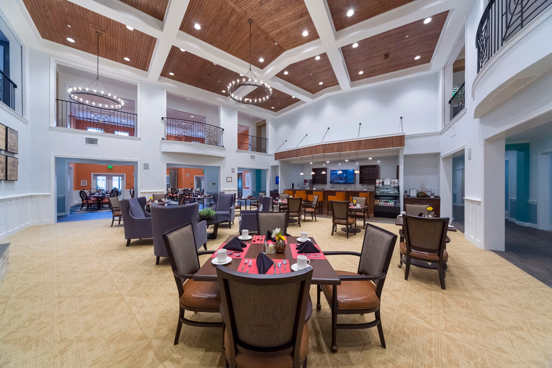 Spacious and well-lit dining area in a senior living facility with several tables set with cups, plates, and napkins. The room features high ceilings with wooden paneling, modern chandeliers, and a balcony railing on the upper level. There is a counter area with a television and beverage coolers in the background.