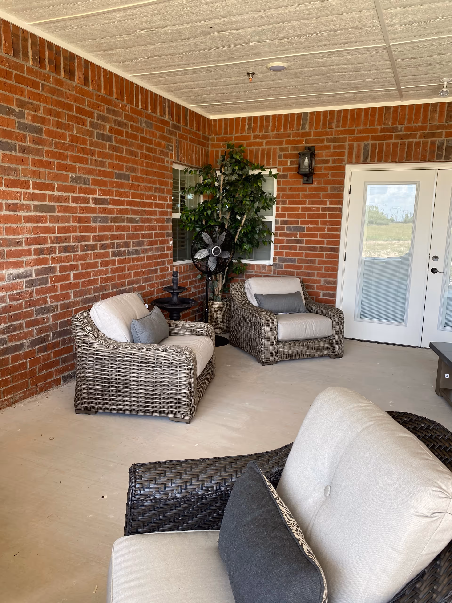 Covered outdoor patio area with brick walls, featuring three cushioned wicker armchairs with pillows, a potted artificial plant, a standing fan, a small black side table, and double glass doors leading inside.