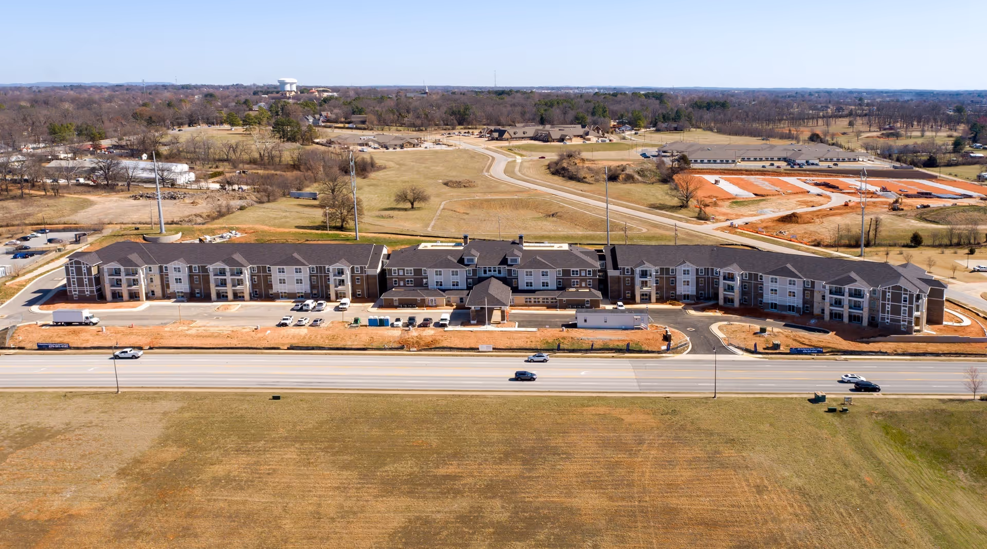 Aerial view of Pinnacle Springs Retirement Community showing a large, multi-wing building with a dark roof and light-colored exterior walls. The building is surrounded by open grassy areas, a few trees, and a road with several cars driving by. In the background, there are more buildings, trees, and a water tower under a clear blue sky.