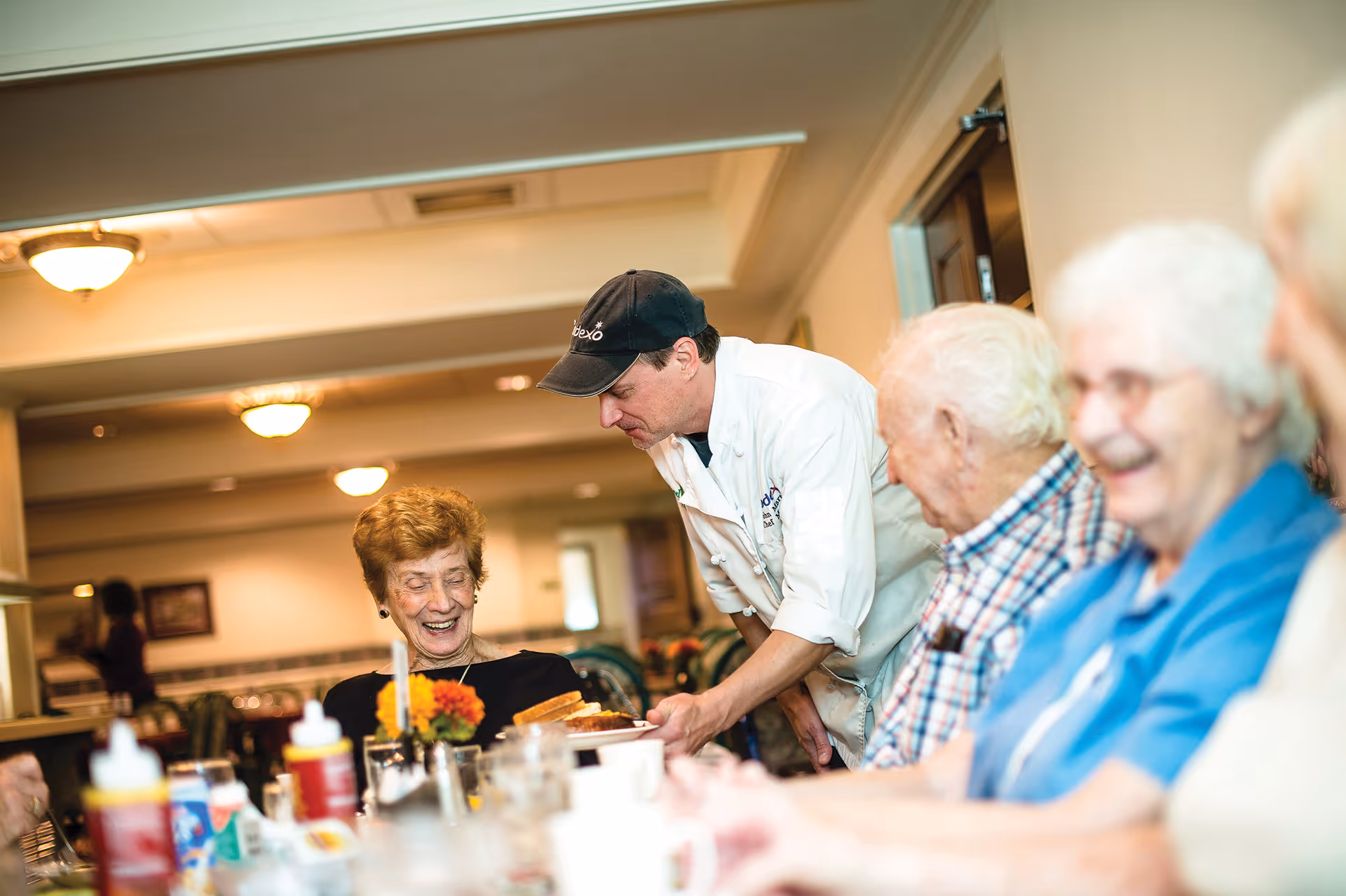 A chef serving food to a smiling elderly woman seated at a dining table with other elderly people in a well-lit dining room.