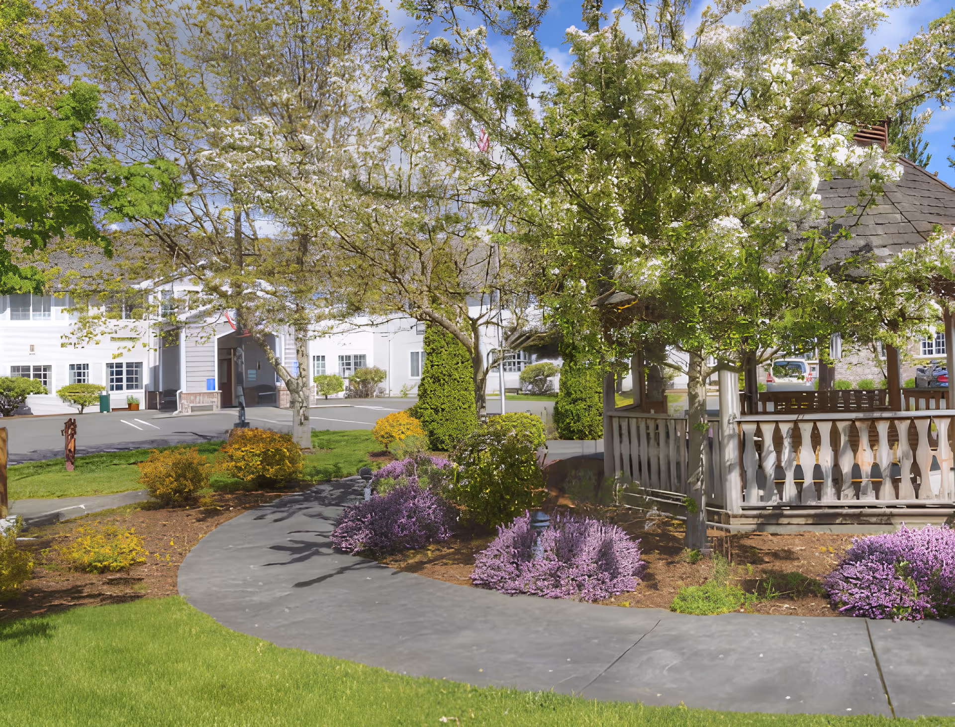 A landscaped outdoor area at Trustwell Living at Sinclair Place featuring a curved paved walkway, blooming trees with white flowers, purple and yellow shrubs, a wooden gazebo with a shingled roof, and a white building with multiple windows in the background under a partly cloudy sky.