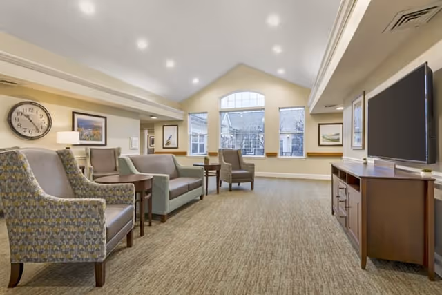 A spacious and well-lit living room area in Brookdale Pittsford featuring comfortable armchairs and sofas arranged around small wooden tables. The room has a large window with an arched top letting in natural light, beige walls adorned with framed artwork, a wall clock, and a large flat-screen TV mounted on a wooden media console.