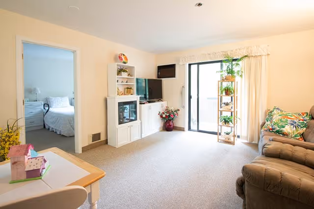 Bright living room with a reclining sofa, TV cabinet and shelving, a sliding glass door to a balcony, and an open doorway showing a bedroom.