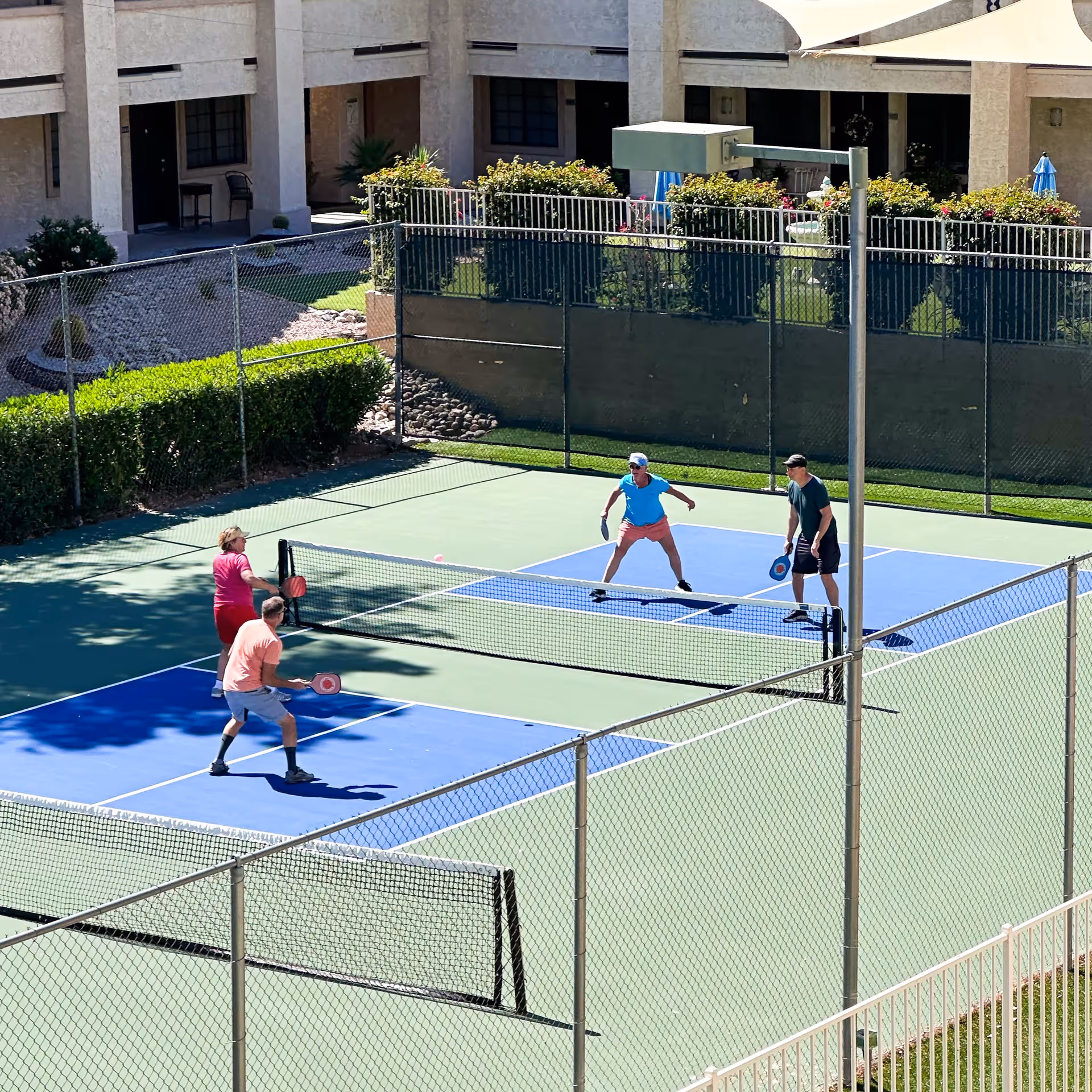 Four people playing pickleball on an outdoor court surrounded by a chain-link fence, with a senior living facility building and landscaped garden in the background.