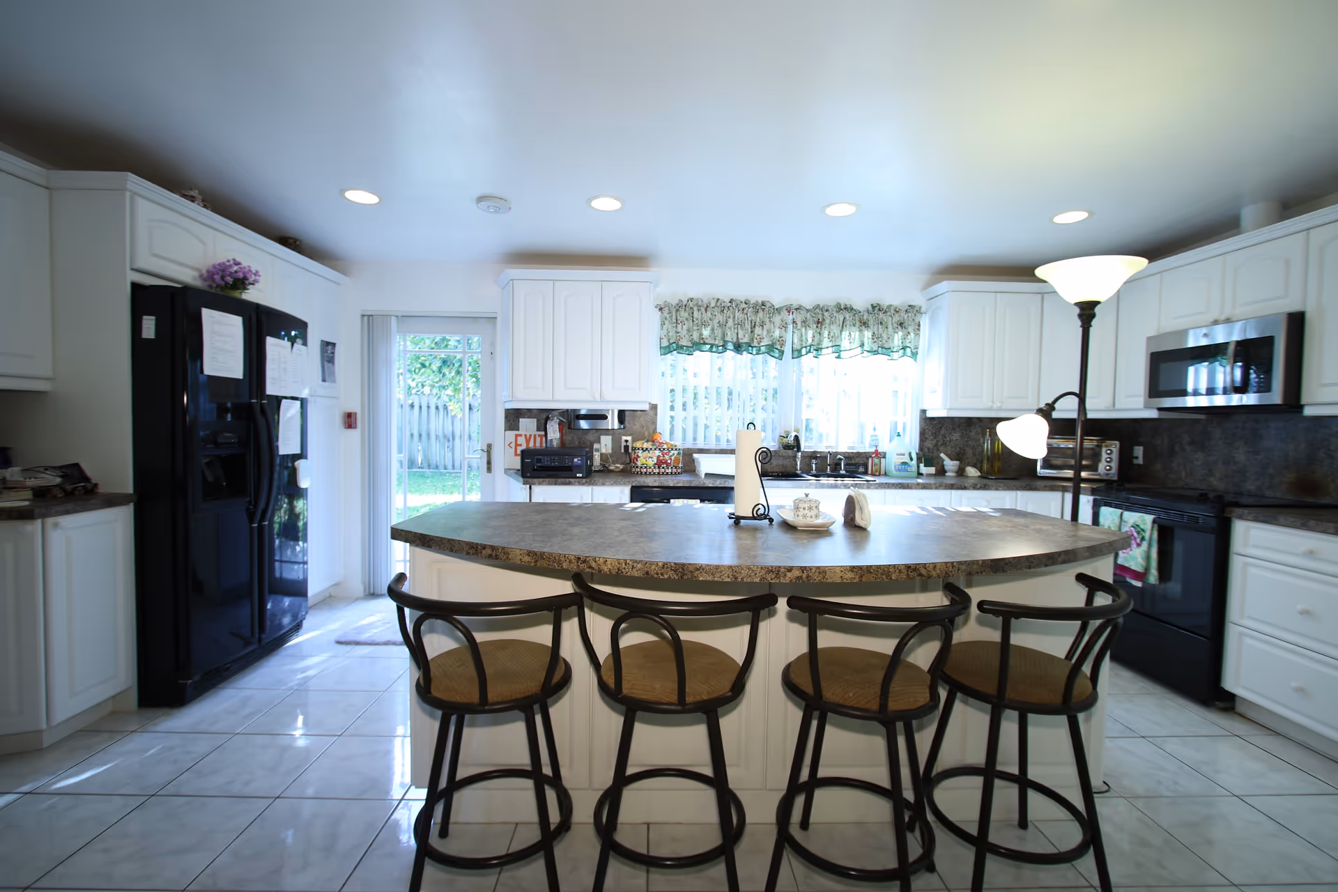 Bright kitchen with white cabinets, a black refrigerator, a black stove, a microwave, and a large island with four bar stools. There is a floor lamp next to the island, a window with floral curtains above the sink, and a glass door leading outside.