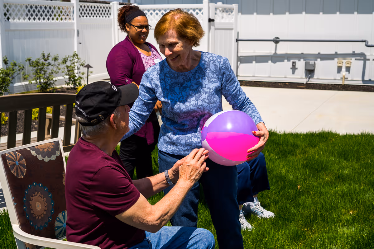 Three seniors enjoying time outdoors on a sunny day. One woman in a blue patterned shirt is holding a purple and pink beach ball and smiling at a seated man wearing a black cap and maroon shirt. Another woman in a purple cardigan stands in the background near a white fence.