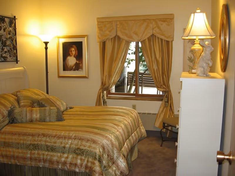A cozy bedroom with a bed covered in a striped gold and green comforter and matching pillows. There is a floor lamp and a framed portrait of a young girl on the wall to the left. A window with gold curtains tied back reveals a view of an outdoor bench and greenery. On the right side, there is a white dresser with a lamp and decorative items on top, and a chair partially visible beside it.