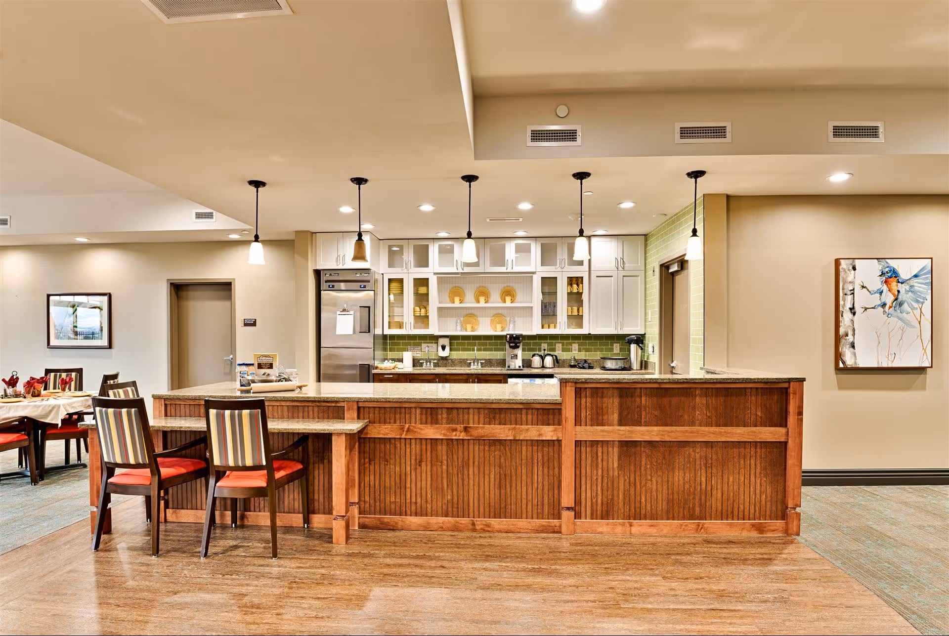 A modern kitchen area in a senior living facility with a wooden counter and bar stools with striped cushions. The kitchen has white cabinets, a stainless steel refrigerator, green tiled backsplash, and hanging pendant lights. To the left, there is a dining area with tables and chairs, and on the right wall, there is a framed painting of a bird.