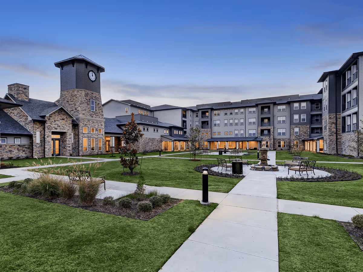 Courtyard of a senior living complex featuring a clock tower, central fountain, benches, walkways, and surrounding multi-story buildings at dusk.