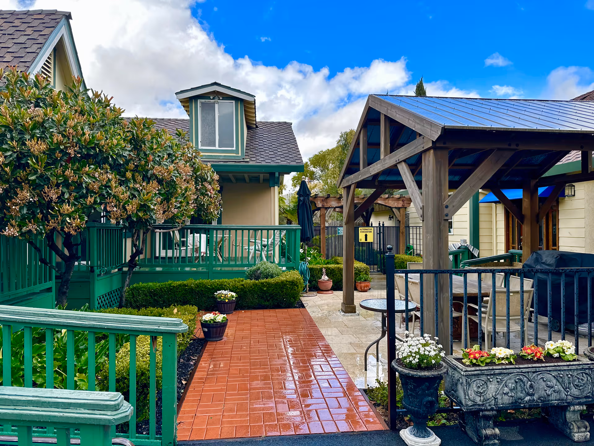 A landscaped outdoor courtyard with a red brick walkway, green railings, potted flowers, and a wooden pergola beside residential buildings.