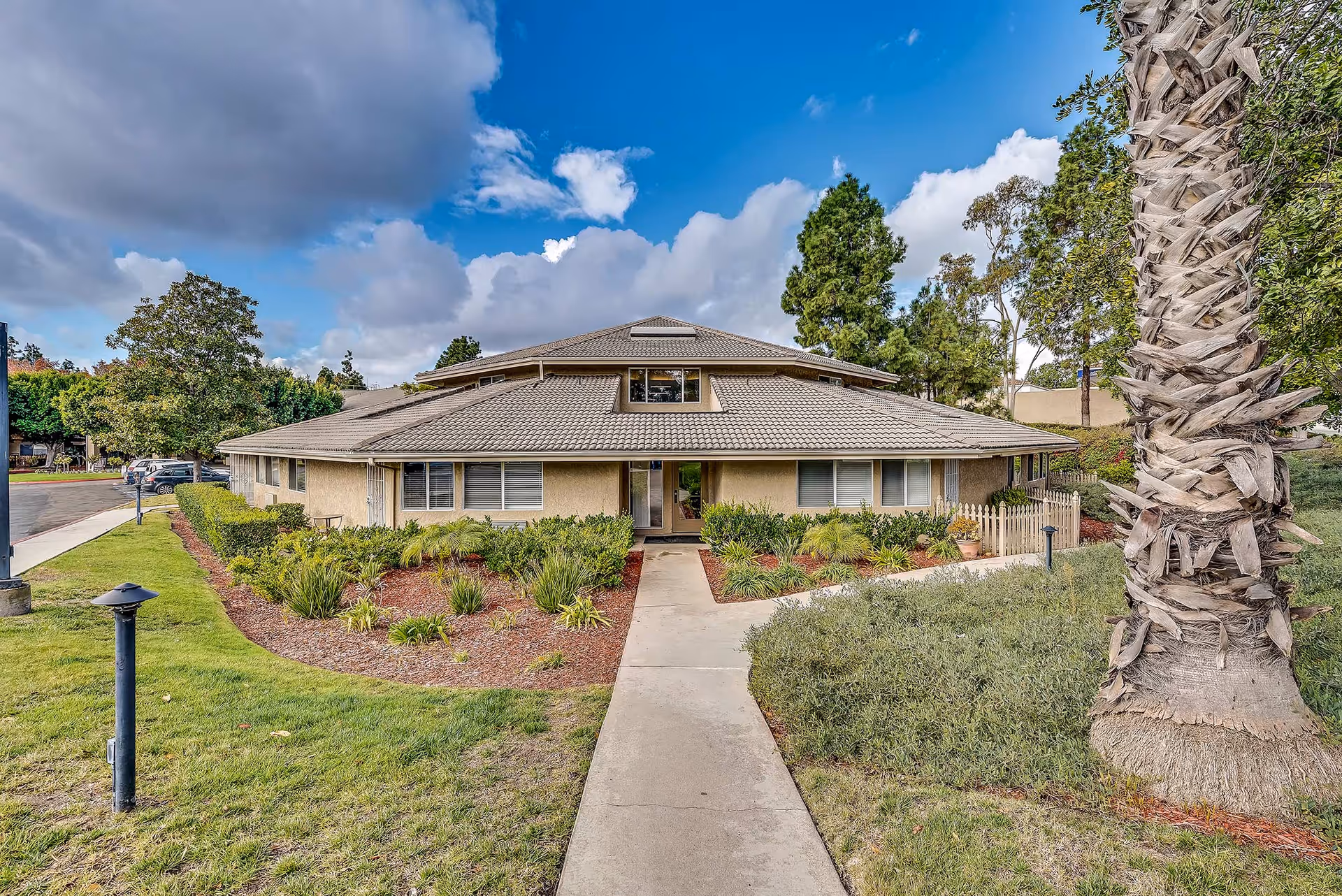 Front exterior view of a single-story building with a tiled roof, surrounded by landscaped greenery including bushes, trees, and a palm tree. A concrete walkway leads to the entrance under a partly cloudy blue sky.