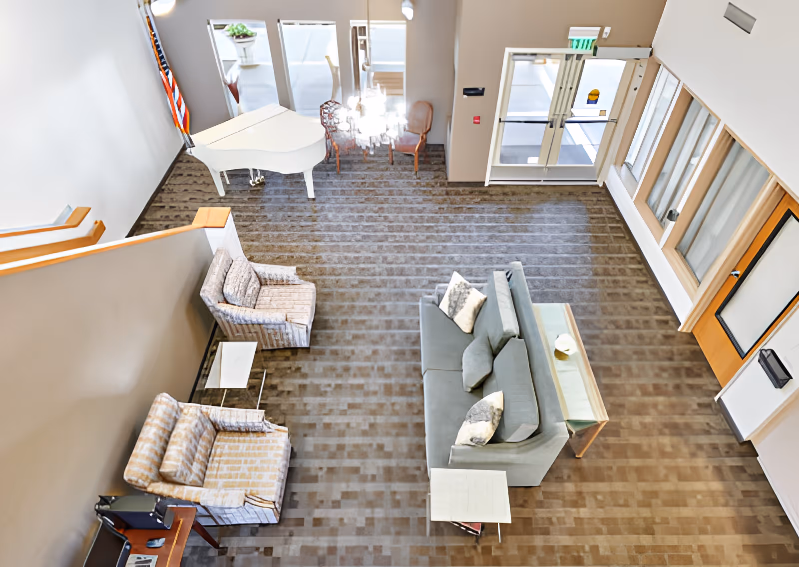 A spacious living room area viewed from above, featuring a gray sofa with pillows, two patterned armchairs, a white grand piano near the wall with an American flag, a glass coffee table, and a chandelier hanging above a small dining table with chairs near the entrance doors.