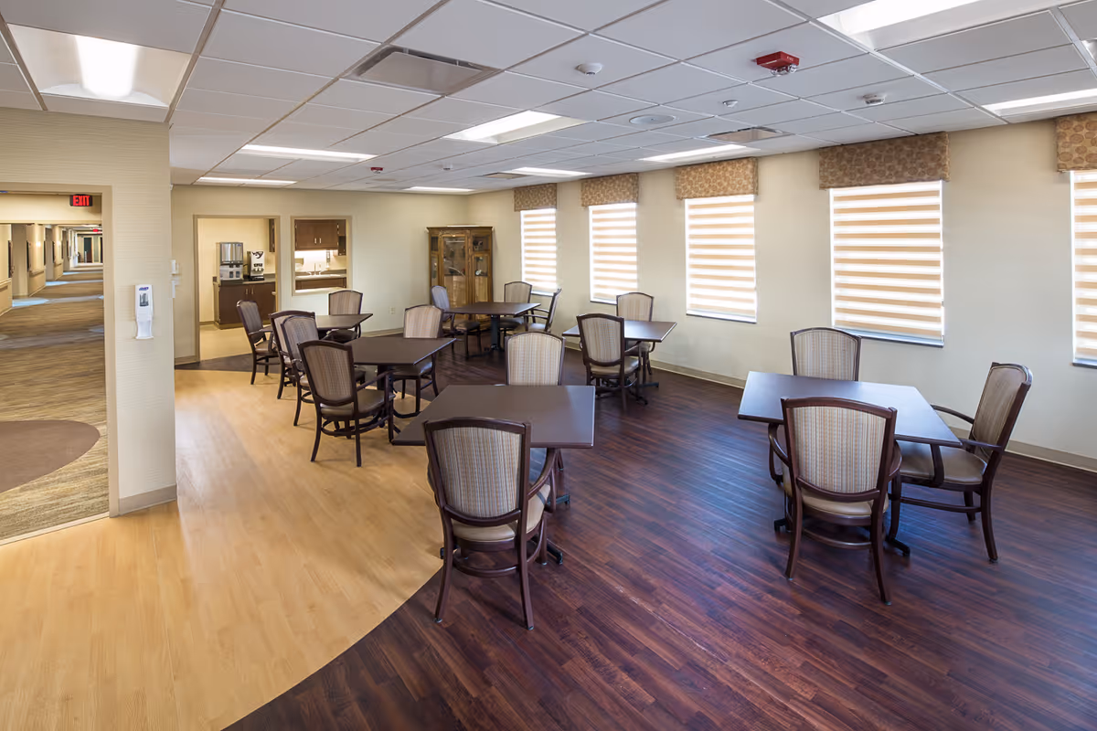 A dining area in a senior living facility with several square tables and cushioned chairs arranged neatly. The room has large windows with striped blinds allowing natural light to enter. The floor features a combination of light and dark wood finishes. In the background, there is a hallway and a small kitchenette area with a coffee machine and cabinets.