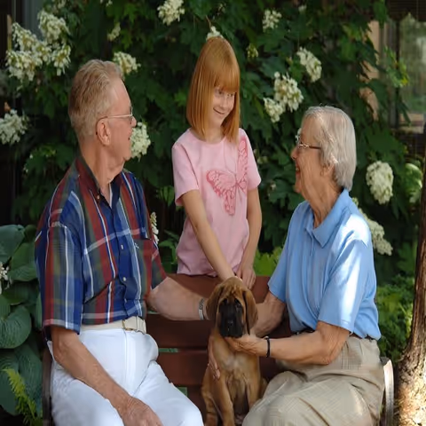 An elderly man and woman sitting on a bench outdoors with a young girl standing between them. They are petting a brown puppy. There are green plants and white flowers in the background.