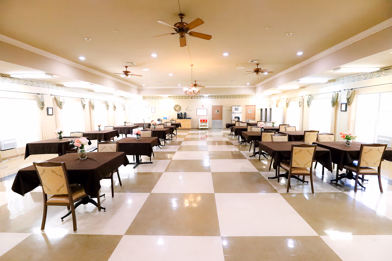 A spacious dining room with multiple tables covered in dark brown tablecloths, each adorned with a small flower arrangement. The room has a checkered floor with beige and white tiles, ceiling fans, and large windows with curtains allowing natural light to fill the space.