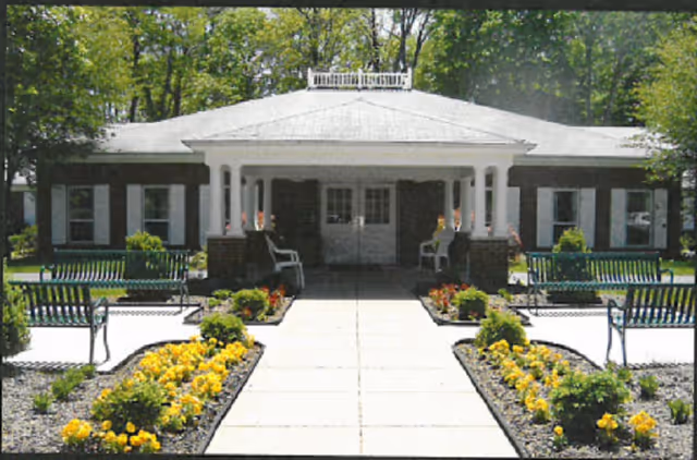 Front exterior view of a single-story building with a white roof and brick pillars supporting a covered entrance. The walkway leading to the entrance is flanked by flower beds with yellow flowers and green shrubs, and there are green benches on either side of the path. Trees are visible in the background.