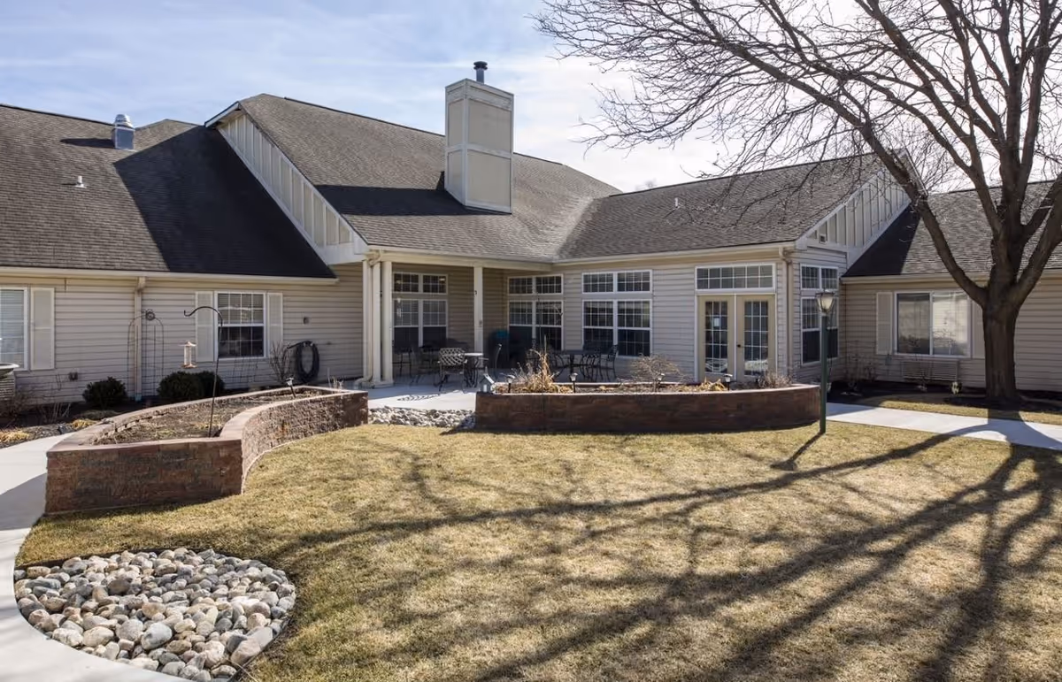 Outdoor view of a single-story assisted living facility with beige siding, multiple windows, and a patio area with tables and chairs. There are raised garden beds, a tree casting shadows on the lawn, and a stone-lined circular area in the grass.