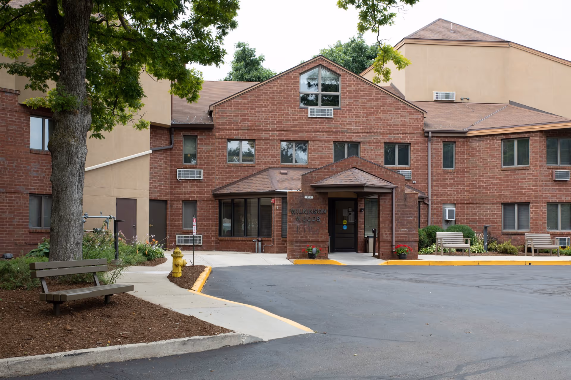 Front exterior view of Wilkinson Woods Senior Community building with brick facade, multiple windows, a main entrance with a small covered porch, benches, a fire hydrant, and trees surrounding the area.