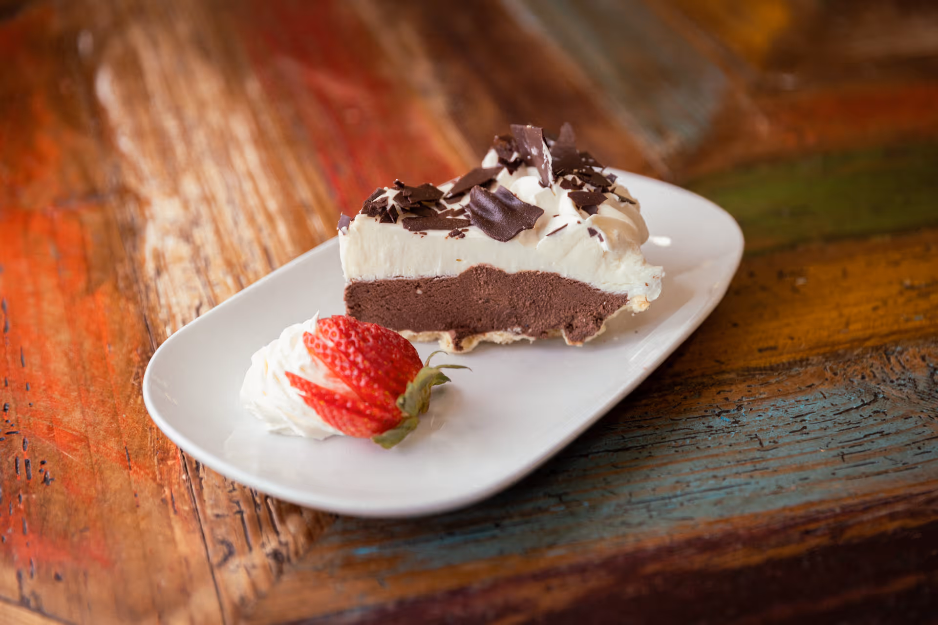 A slice of chocolate cream pie topped with whipped cream and chocolate shavings on a white plate with a fan-cut strawberry garnish on a rustic wooden table.