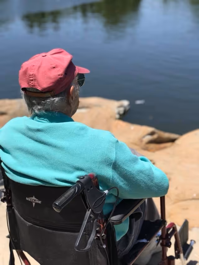 An elderly person wearing a red cap and teal sweater sitting in a wheelchair by a body of water with rocks along the shore, viewed from behind.