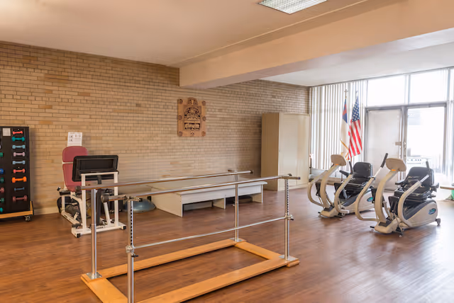 A rehabilitation or exercise room with wooden flooring, featuring parallel bars for walking exercises in the center. To the left, there is a leg exercise machine and a rack with colorful dumbbells. On the right side, there are two recumbent exercise bikes near large windows with vertical blinds. The room has beige brick walls, a wooden plaque on the wall, and two flags near the windows.