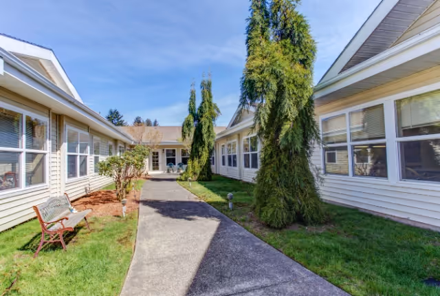 Outdoor courtyard area between two single-story buildings with white siding and multiple windows. The courtyard features a paved walkway, green grass, several tall evergreen trees, a bench, and some small shrubs under a clear blue sky.