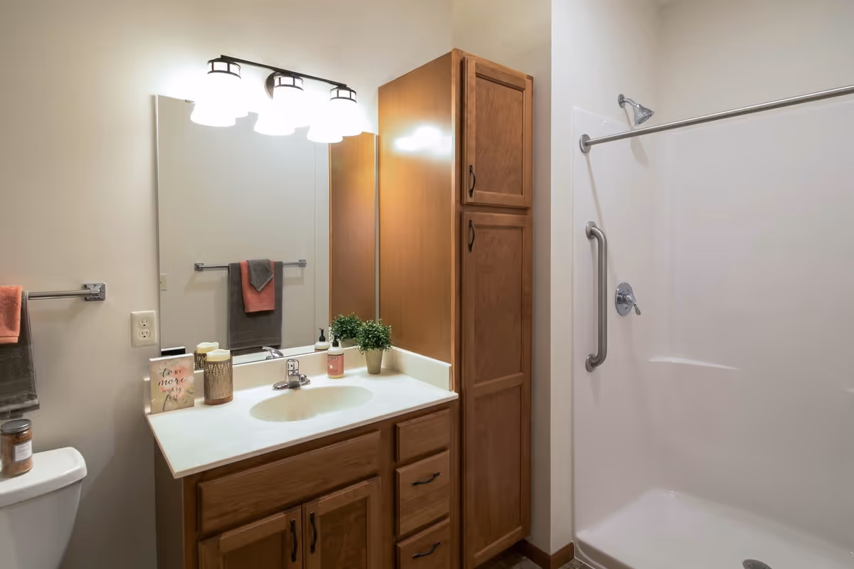 A bathroom with a wooden vanity featuring a white countertop and built-in sink. Above the sink is a large mirror with three light fixtures. To the right is a tall wooden cabinet and a shower area with a grab bar and showerhead. On the left, there is a toilet with a candle on top and two towel racks holding gray and pink towels.