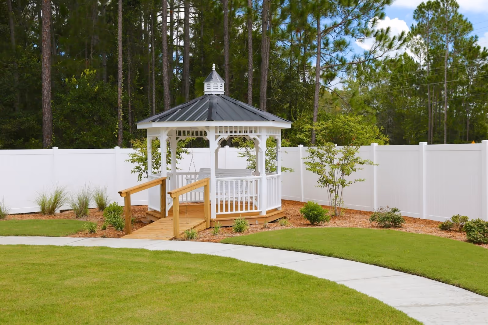 A white gazebo with a black roof and wooden ramp is situated in a landscaped garden area with green grass, small shrubs, and trees. The gazebo is enclosed by a white fence, and tall pine trees are visible in the background under a partly cloudy sky.