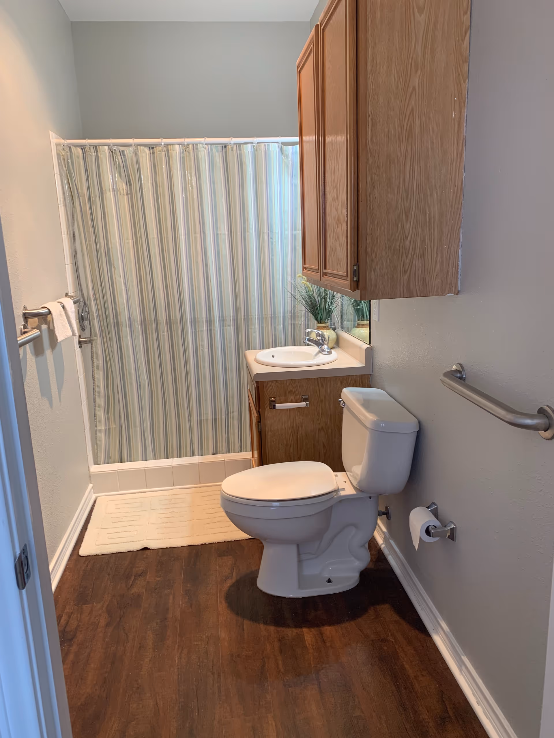A bathroom with a toilet, a sink with a wooden cabinet above and below it, a striped shower curtain, a towel rack with a white towel, a grab bar on the wall, and a beige bath mat on a dark wood floor.