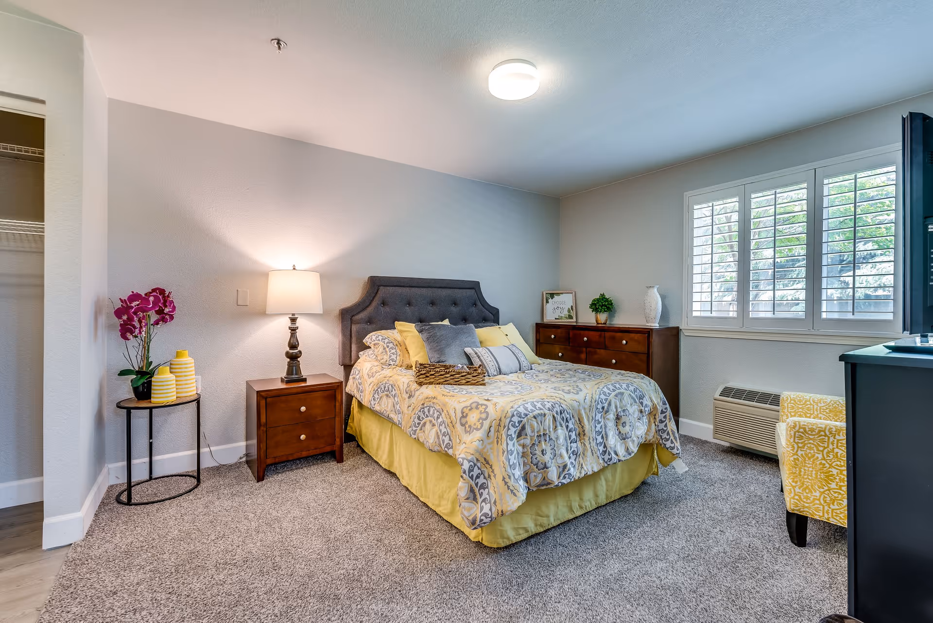 A cozy bedroom with a bed featuring a patterned yellow and gray bedspread, a dark upholstered headboard, and multiple pillows. To the left of the bed is a wooden nightstand with a lamp and a small round table with yellow vases and a purple orchid. On the right side of the room is a wooden dresser with decorative items and a window with white shutters letting in natural light. A yellow patterned armchair is partially visible near the window, and the floor is carpeted.