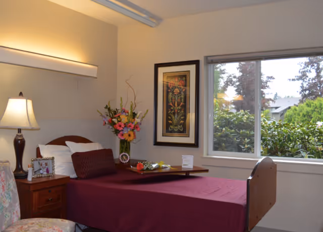 A cozy bedroom in a rehabilitation health center with a single bed covered in a maroon bedspread, a bedside table with a lamp and framed photo, a floral arrangement on a tray on the bed, a framed artwork on the wall, and a window showing greenery outside.